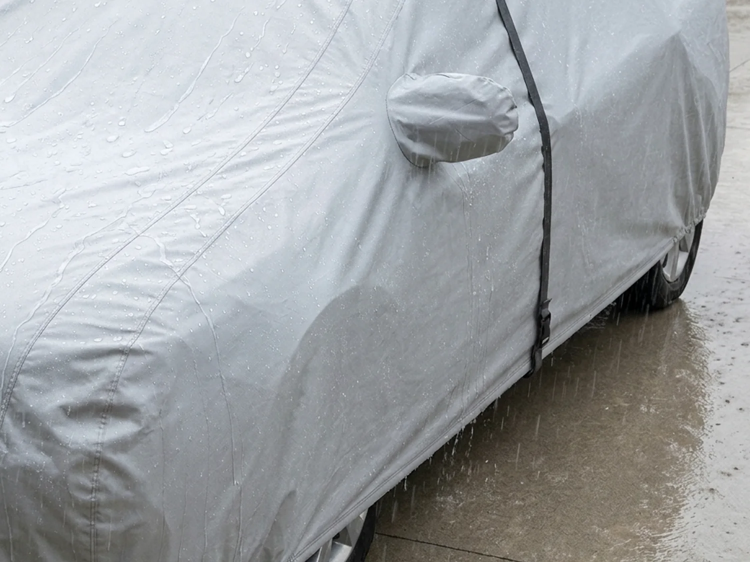 Heavy rain beading and running off the silver-grey cover fitted over a sedan on a driveway