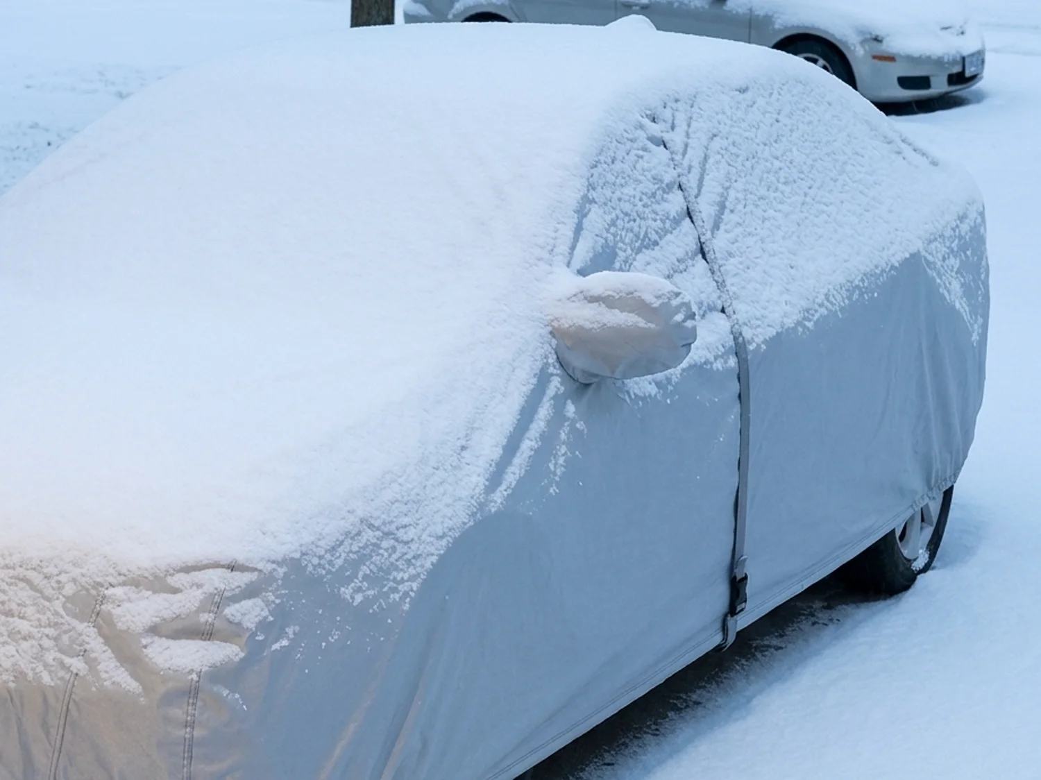 A covered sedan under a layer of snow on a driveway with an uncovered car behind it